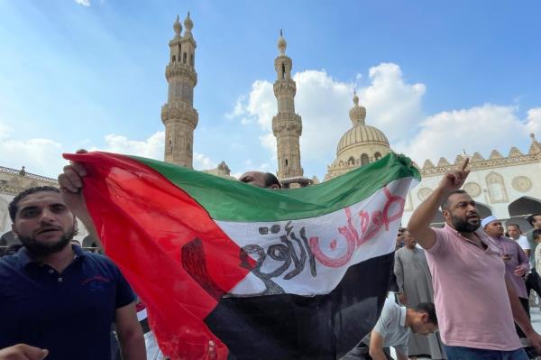 A group of protestors holding a Palestinian flag with Arabic writing on it in a large courtyard mosque with minarets rising above in the background.