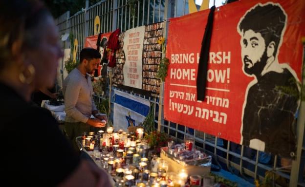 A row of candles sits in front of posters depicting hostages. One poster says 'Bring Hersh home now!' with a drawing of a man. 